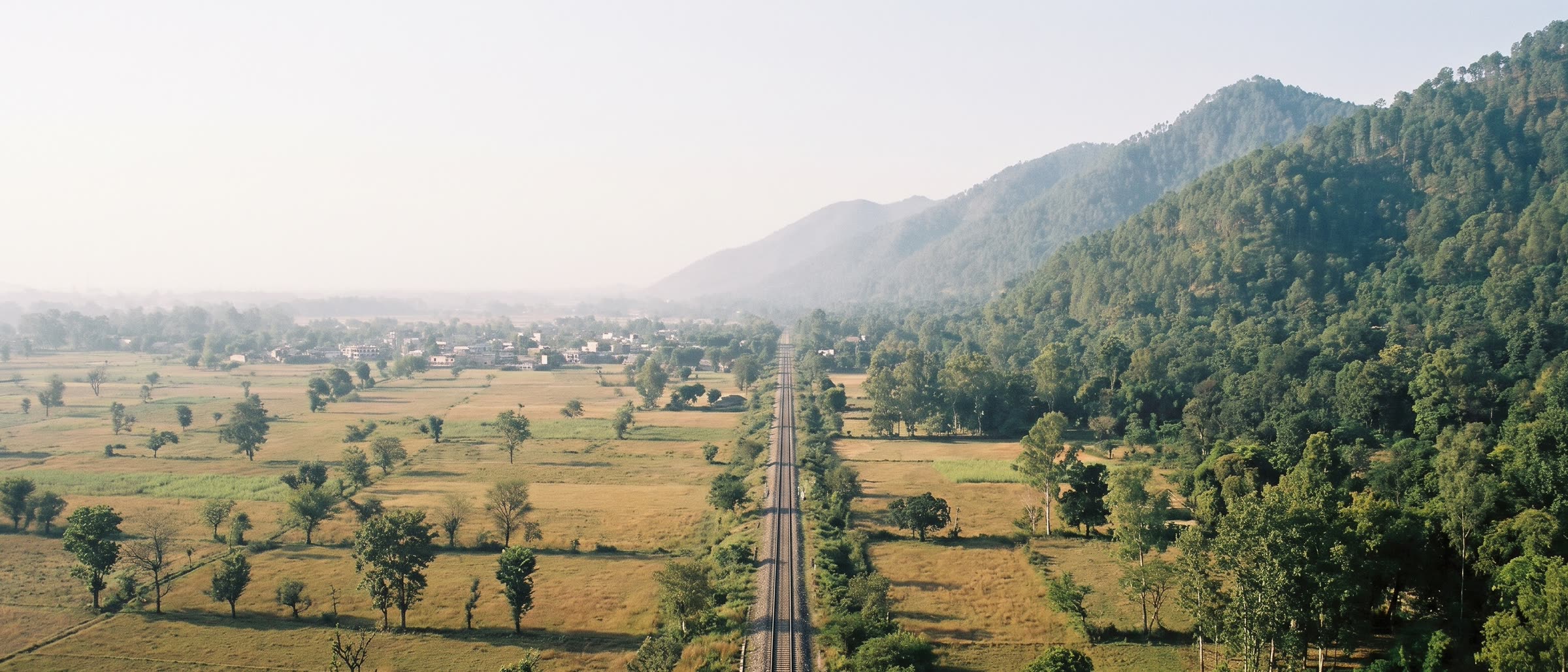 A flat plain of fields and trees, a railway line running straight across it to the foot of a steep forested ridge.