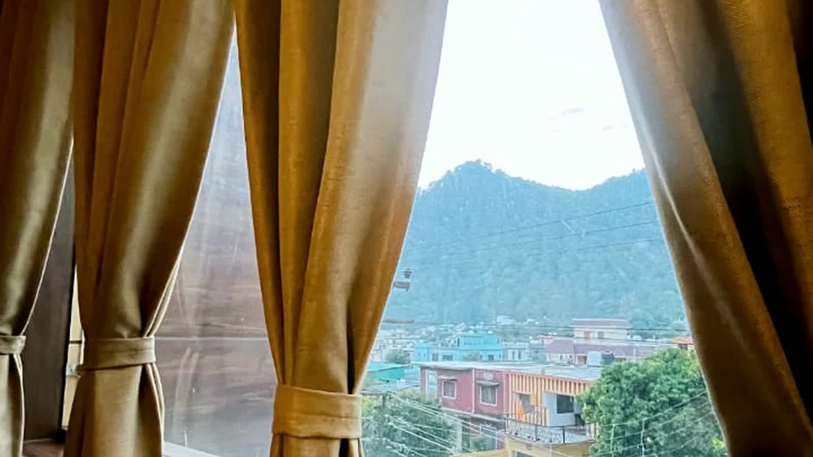Gold curtains drawn back from a guest-room window, rooftops below and a forested ridge behind.